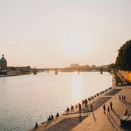 Sonnenuntergang an der Garonne in Toulouse, an deren Uferpromenade gemächlich Menschen spazieren und sitzen. Die Silhouette einer Kuppel und einer Brücke in der Ferne unterstreicht die friedliche Szenerie.