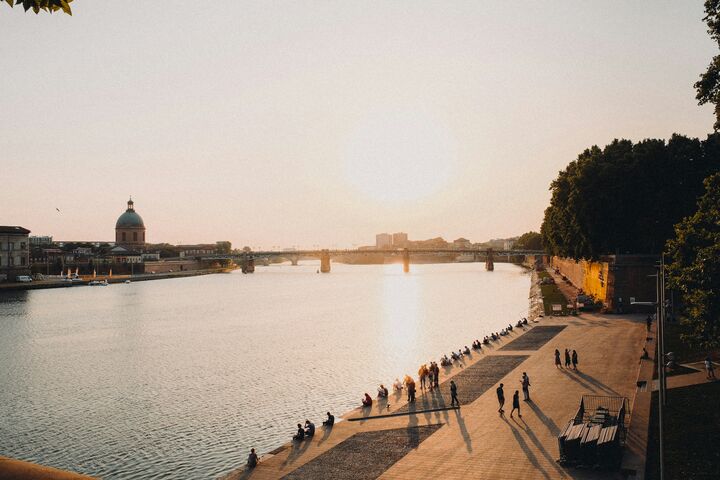 Sonnenuntergang an der Garonne in Toulouse, an deren Uferpromenade gemächlich Menschen spazieren und sitzen. Die Silhouette einer Kuppel und einer Brücke in der Ferne unterstreicht die friedliche Szenerie.