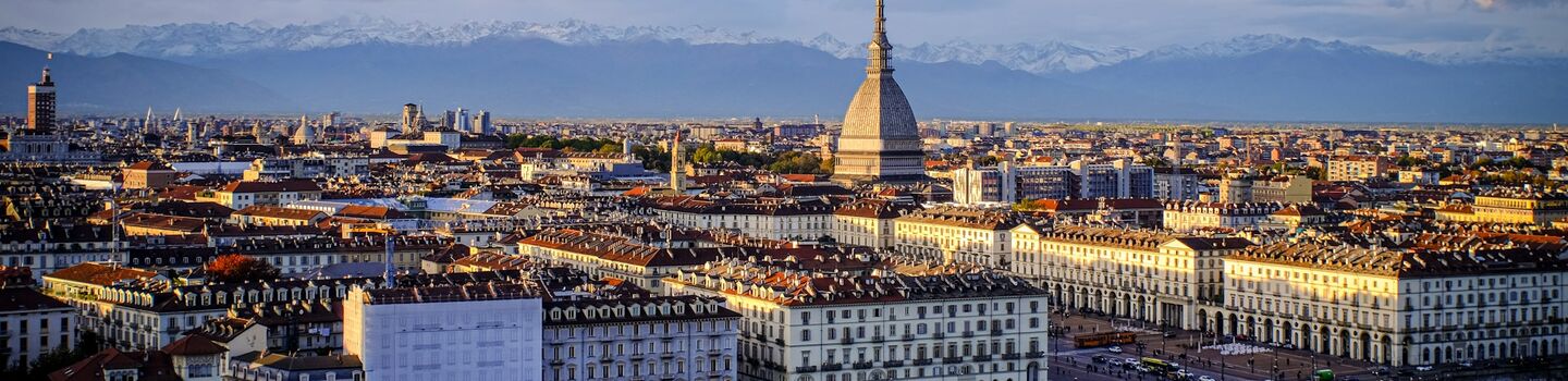 Luftaufnahme von Turin, mit der ikonischen Mole Antonelliana, die die Stadt überragt. Sonnenbeschienene Gebäude, üppige Bäume und in der Ferne schneebedeckte Alpen unter blauem Himmel.