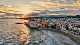 Luftaufnahme der Küstenstadt Sitges bei Sonnenuntergang: Bunte Gebäude säumen das Ufer, während goldenes Licht über das Meer schimmert und eine friedliche Stimmung schafft.