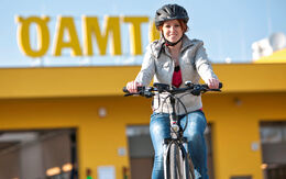 Radfahrerin auf Fahrrad mit beiger Jacke, Jeans und schwarzen Radhelm fährt vor ÖAMTC-Stützpunk-Gebäude.