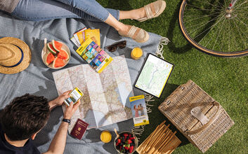 Zwei Personen sitzen auf einer Picknickdecke im Gras. Auf der Decke liegen zudem Landkarten, ein Tablet, Reiseführer, Obst, ein Hut. Im Eck sieht man den Reifen eines Fahrrads.