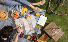 Zwei Personen sitzen auf einer Picknickdecke im Gras. Auf der Decke liegen zudem Landkarten, ein Tablet, Reiseführer, Obst, ein Hut. Im Eck sieht man den Reifen eines Fahrrads.