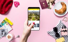Eine Hand hält ein Smartphone mit einem Bild von Paris. Im pinkfärbigen Hintergrund liegen ein Reiseführer, ein Reisepass, ein Croissant, eine Sonnenbrille, ein Herz-Macchiato, eine rote Baskenmütze und weitere Reiseutensilien.
