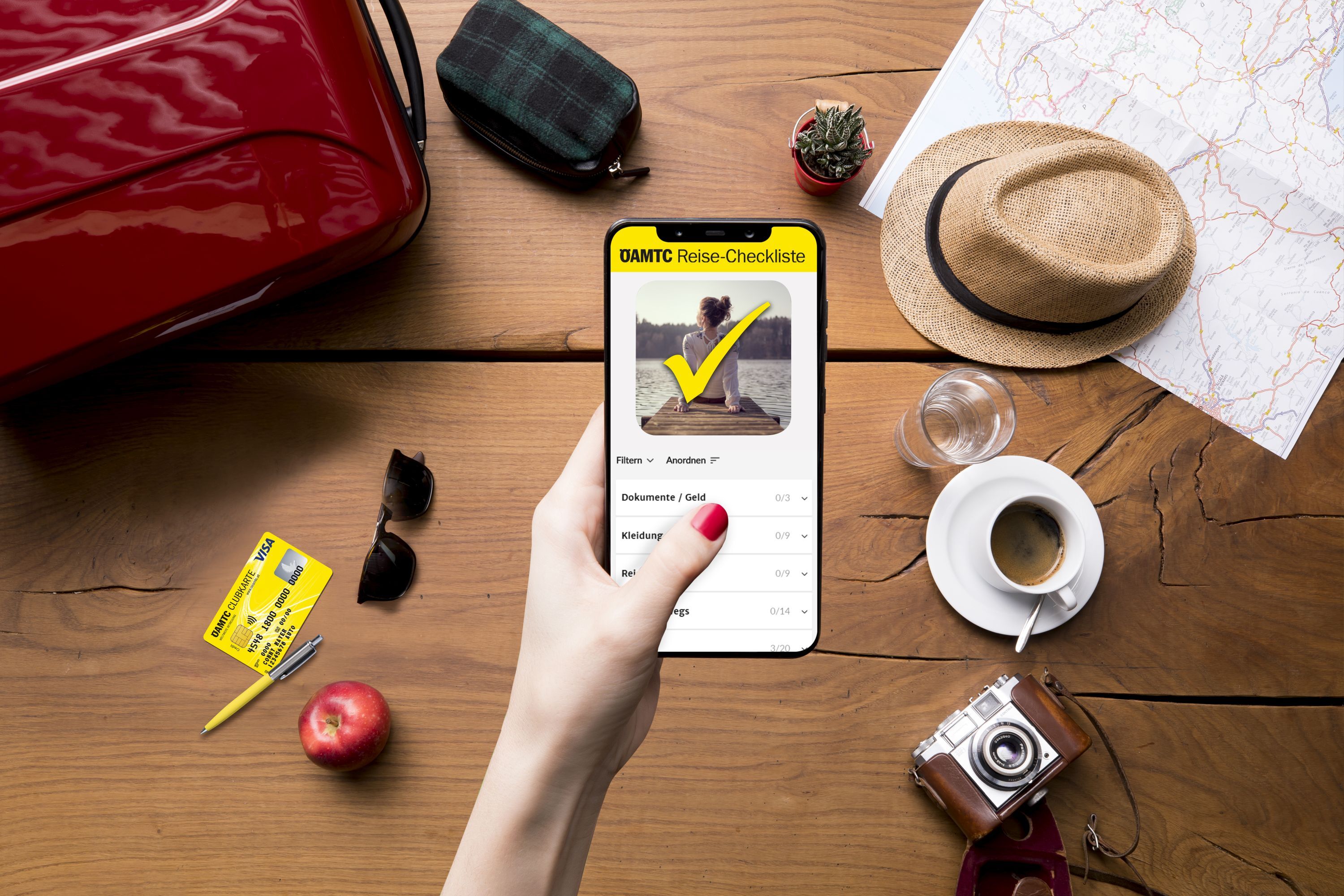 Eine Hand hält ein Smartphone mit der ÖAMTC Reise-Checkliste, umgeben von Reisetasche, Hut, Karte, Kamera, Kaffee und Apfel.