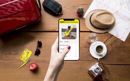 Eine Hand hält ein Smartphone mit der ÖAMTC Reise-Checkliste, umgeben von Reisetasche, Hut, Karte, Kamera, Kaffee und Apfel.
