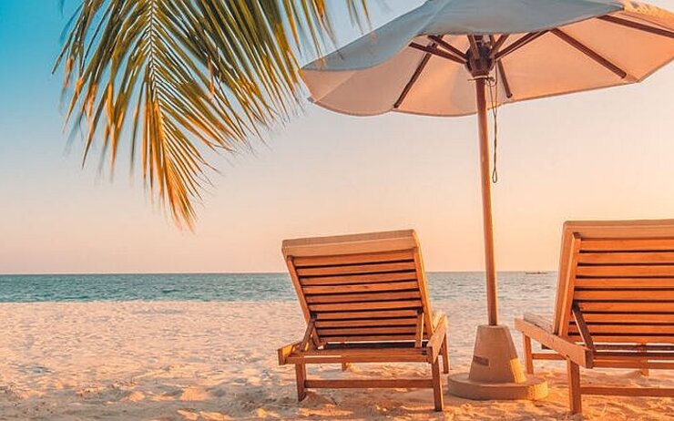Zwei Liegestühle aus Holz und in der Mitte ein Sonnenschirm stehen am Sandstrand. Ansicht von hinten.