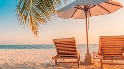 Zwei Liegestühle aus Holz und in der Mitte ein Sonnenschirm stehen am Sandstrand. Ansicht von hinten.