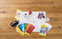 Karte auf Holztisch, umgeben von Reiseführern, Apfel, Reisepass, Sonnenbrille, Tasse und Kamera.