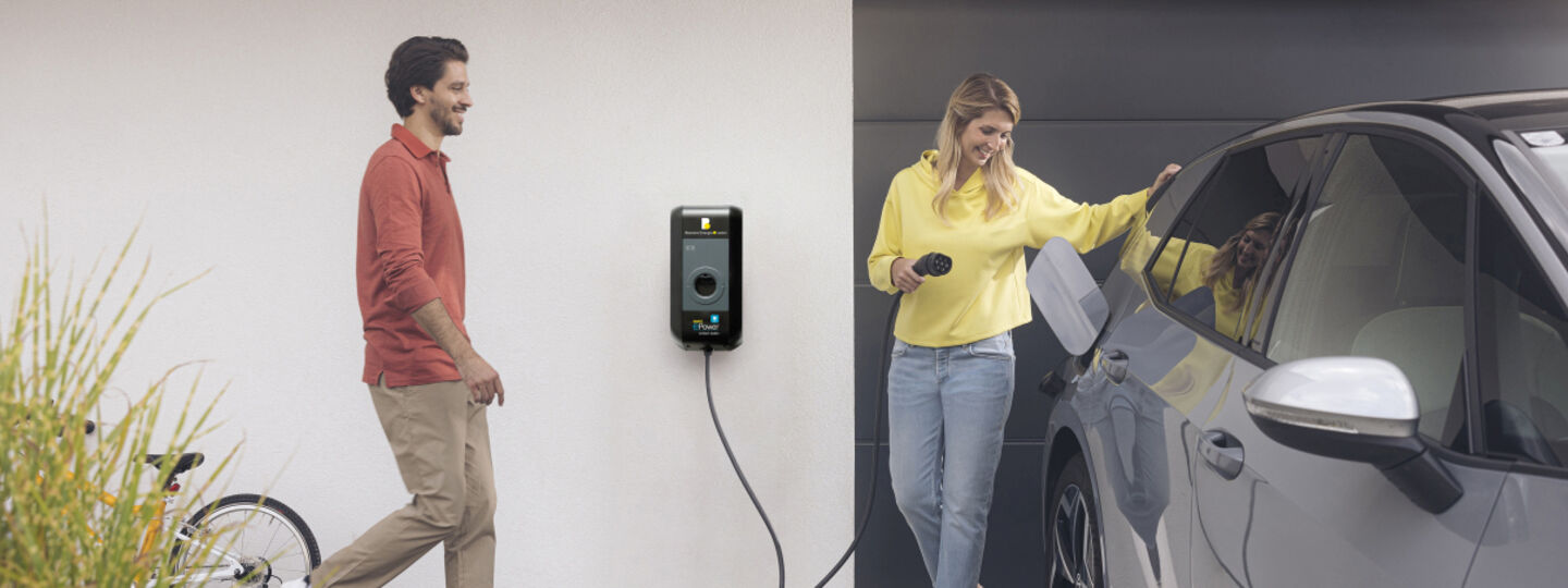Eine Frau lädt ein Elektroauto vor einem Haus mit eigener Wallbox, ein Mann steht neben ihr.