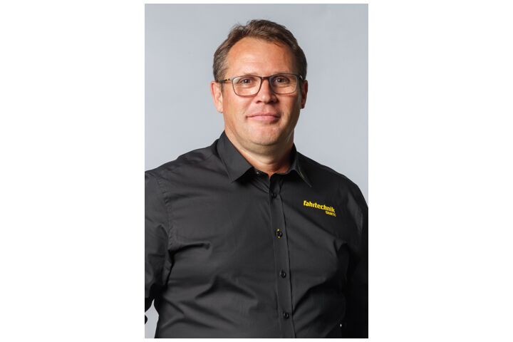 Ein Mann mit Brille und schwarzem Hemd, mit der Aufschrift Fahrtechnik ÖAMTC, steht vor einem hellgrauen Hintergrund und blickt zufrieden.