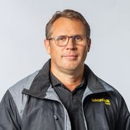 Das Bild ist ein Portrait von Roland Frisch, der frontal in die Kamera schaut und eine schwarze Jacke mit gelber ÖAMTC Fahrtechnik-Aufschrift trägt.