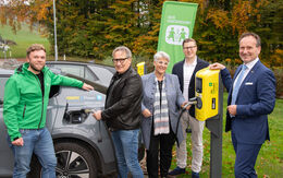 Vor einem Elektroauto stehen fünf Personen, ein Mann hält das Ladekabel beim Auto, eine Frau hält es an der Ladestation. Am Auto steht ÖAMTC ePower - einfach laden. Hinter den Personen ist eine grüne Fahne mit der Aufschrift SOS Kinderdorf. Im Hintergrund Wiesen und Bäume.