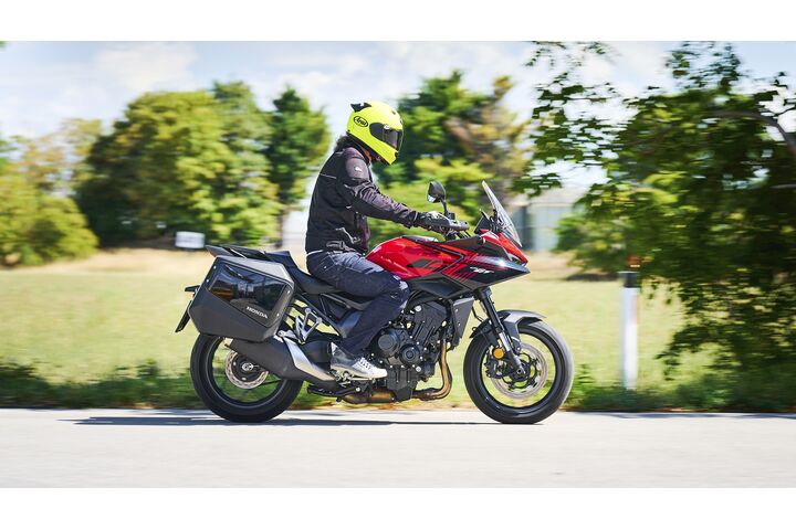 Motorradfahrerin oder Motorradfahrer mit gelbem Helm fährt eine rote Honda CB1000GT mit schwarzen Seitenkoffern auf einer Landstraße; der Hintergrund ist durch die Bewegung unscharf.