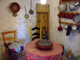 Auf dem Foto ist das Wohnzimmer in einer kleinen Felsenwohnung in Sacromonte bei Grananda zu sehen - runder Tisch, Sessel und Alltagsgegenstände an der Wand. 