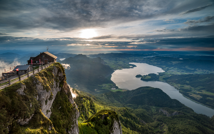 Ein schöner Ausblick von einem Berggipfel, mit einem kleinen Gebäude am Rand, Blick auf einen See und umliegende grüne Landschaften bei Sonnenuntergang.