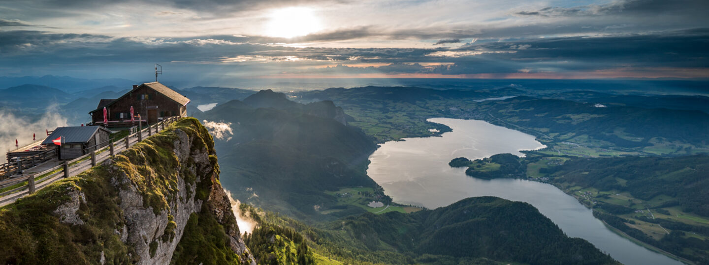 Ein schöner Ausblick von einem Berggipfel, mit einem kleinen Gebäude am Rand, Blick auf einen See und umliegende grüne Landschaften bei Sonnenuntergang.