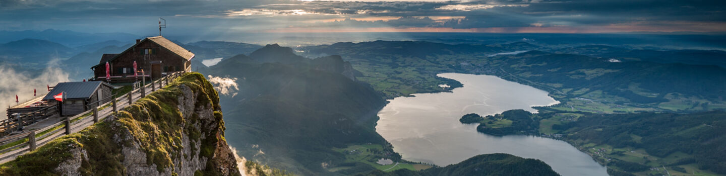 Ein schöner Ausblick von einem Berggipfel, mit einem kleinen Gebäude am Rand, Blick auf einen See und umliegende grüne Landschaften bei Sonnenuntergang.