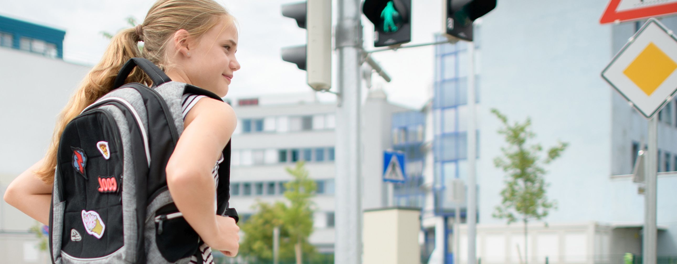 Ein Mädchen steht an einer Ampel, sie trägt einen schwarz-grauen Rucksack und ist seitlich von hinten zu sehen. Im Hintergrund sind eine Fußgängerampel und eine Kreuzung mit großen Firmengebäuden zu sehen..