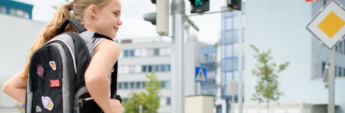 Ein Mädchen steht an einer Ampel, sie trägt einen schwarz-grauen Rucksack und ist seitlich von hinten zu sehen. Im Hintergrund sind eine Fußgängerampel und eine Kreuzung mit großen Firmengebäuden zu sehen..