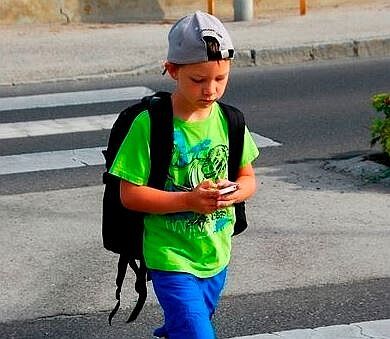 Ein Schulkind mit großem Rucksack am Rücken geht über einen Zebrastreifen. In den Händen hält er ein Handy und blickt darauf.