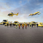 Eine Flotte von gelben Rettungsfahrzeugen und Luftfahrzeugen steht auf einem Flugfeld. Zu sehen ein Krankenwagen, ÖAMTC Pannenfahrzeuge, ein Abschlepp-LKW, ein Rettungshubschrauber in der Luft sowie ein Ambulanzflugzeug. Davor stehen Einsatzkräfte in leuchtender Schutzkleidung. 