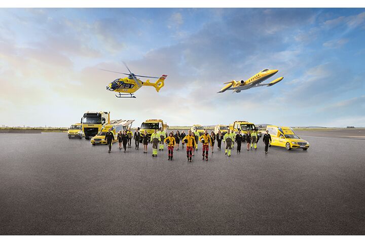 Eine Flotte von gelben Rettungsfahrzeugen und Luftfahrzeugen steht auf einem Flugfeld. Zu sehen ein Krankenwagen, ÖAMTC Pannenfahrzeuge, ein Abschlepp-LKW, ein Rettungshubschrauber in der Luft sowie ein Ambulanzflugzeug. Davor stehen Einsatzkräfte in leuchtender Schutzkleidung. 