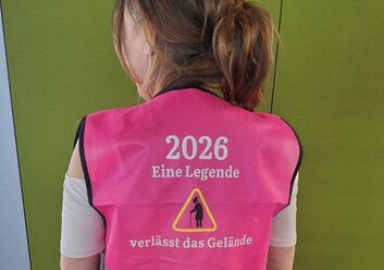 Eine Person steht vor einer grünen Wand. Sie trägt eine rosa warnweste. Auf dieser steht. 2026. Eine Legende verlässt das Gelände
