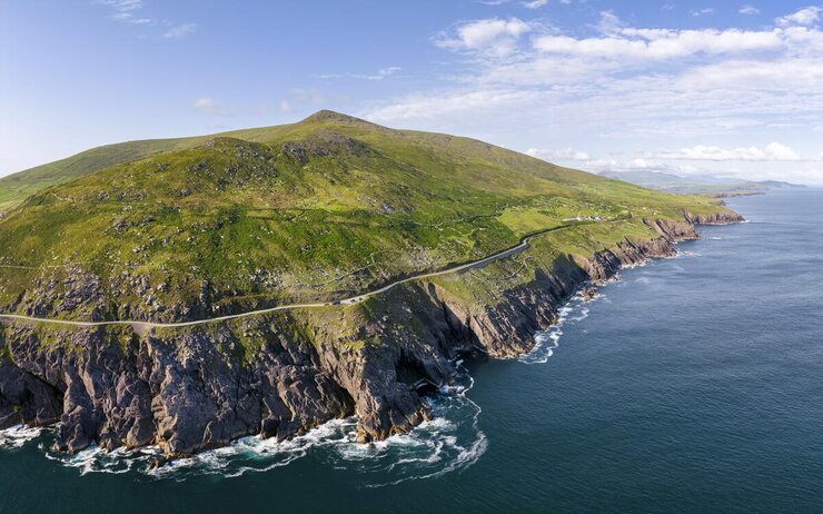 Das Foto zeigt den Slea Head Drive, eine der Panoramaküstenstraßen entlang der schroffen, irischen Felsenküste mit grünen Hüfeln im Hintergrund und dem blauen Meer unterhalb der Felsen.