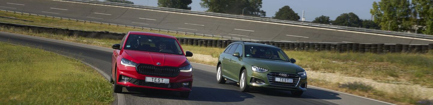 Ein rotes und ein grünes Auto fahren nebeneinander auf einer Teststrecke. Neben der Fahrbahn ist grüne Wiese.