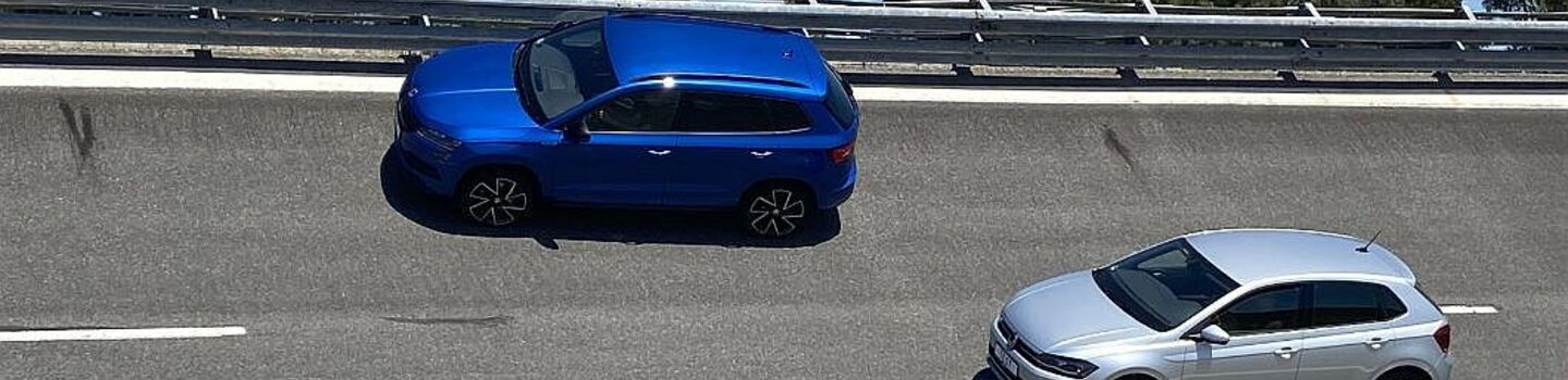 Ein blauer SUV und ein silberner Kleinwagen - Testautos für den Sommerreifentest 2022 - fahren hintereinander auf einer mehrspurigen Straße. Im Hintergrund sind Bäume und ein blauer Himmel zu sehen. 