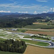 Inmitten einer hügeligen Landschaft erstreckt sich das Fahrtechnik Zentrum St.Veit. Es gibt asphaltierte Trainingsstrecken mit Kreuzungselementen, eine Kreisbahn und Kurvenkombinationen. Im Hintergrund sind Wälder, Häuser und ein See.