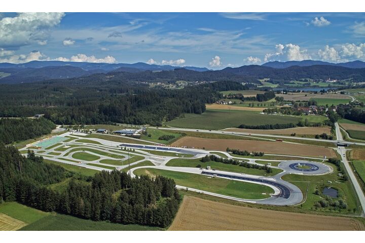 Inmitten einer hügeligen Landschaft erstreckt sich das Fahrtechnik Zentrum St.Veit. Es gibt asphaltierte Trainingsstrecken mit Kreuzungselementen, eine Kreisbahn und Kurvenkombinationen. Im Hintergrund sind Wälder, Häuser und ein See.