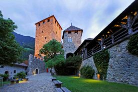 Auf dem Foto ist eine Burgmauer und 2 Türme von Schloss Tirol in Südtirol beleuchtet am Abend zu sehen.