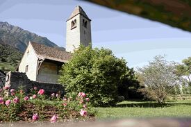 Auf dem Foto ist die St. Prokulus Kirche in Naturns in Südtirol mit blühenden Blumen davor zu sehen.