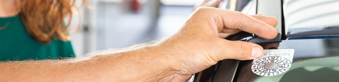 Eine Männerhand, die eine §57a Plakette in die Ecke einer Windschutzscheibe klebt