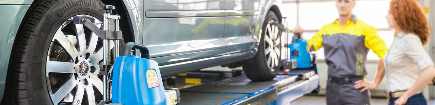 Im Vordergund ist ein blaues Fahrzeug auf einer Hebebühne zu sehen. Im Hintergrund ein ÖAMTC Techniker mit einem weiblichen Mitglied. 