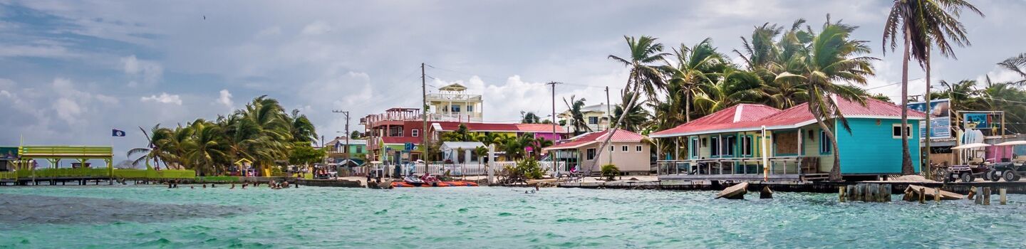 Starker Wind weht durch die Palmen von Belize, wo bunte Häuser am Rande eines graublauen Meeres zu sehen sind.