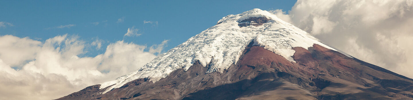 Der Cotopaxi, ein imposanter brauner Berg mit schneeweißer Spitze, zeichnet sich stark ab gegen einen kobaltblauen Himmel, gekrönt von dicken weißen Wolken.