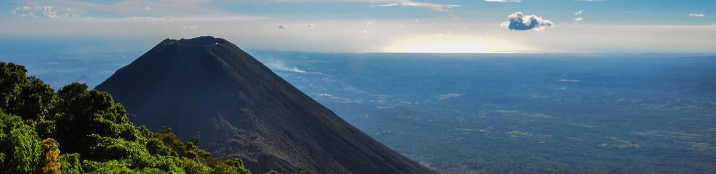 Ein Vulkan ragt majestätisch über einer weiten Ebene, während die letzten Sonnenstrahlen des Tages hinter dem Horizont hervorblitzen.