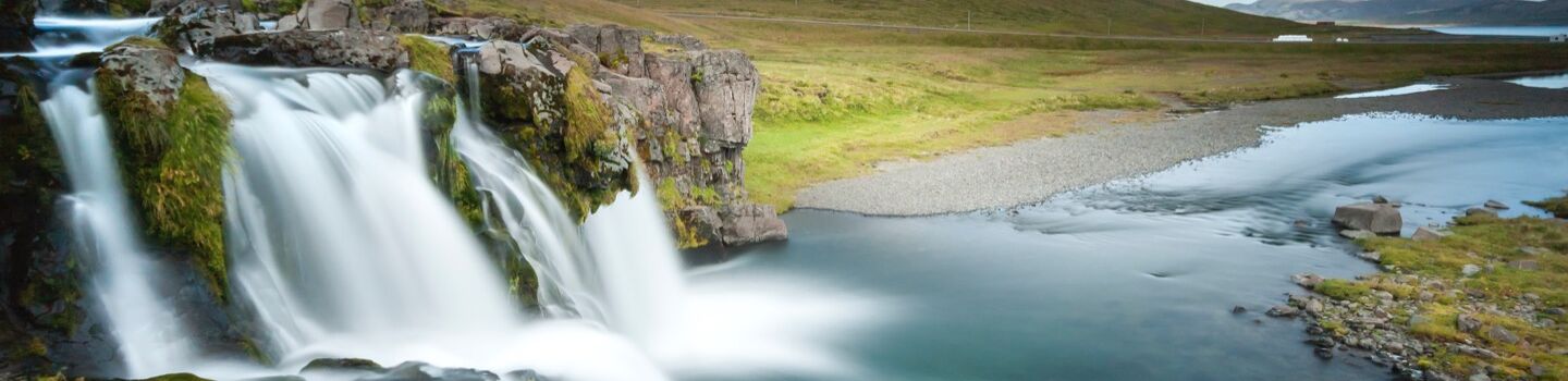 Ein tosender Wasserfall stürzt in einen Fluss, dahinter schroffe Felsen und grüne Hügel