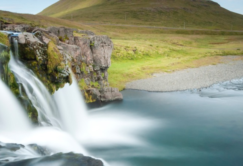 Ein tosender Wasserfall stürzt in einen Fluss, dahinter schroffe Felsen und grüne Hügel