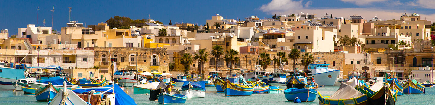 Fischerboote in einem Hafen, im Hintergrund Palmen und alte Steinhäuser an der Hafenpromenade