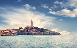 Eine malerische Küstenstadt mit bunten Häusern und einem hohen Kirchturm am Meer unter blauem Himmel.