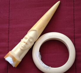 Das Bild zeigt zwei Gegenstände aus Elfenbein auf einem roten Tuch: einen geschnitzten Dolch mit einem Kopf an einem Ende und einen runden Armreif.