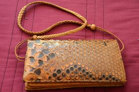 Das Bild zeigt eine kleine Handtasche aus Schlangenleder mit einem geflochtenen Trageriemen, die auf einem roten Tuch liegt.
