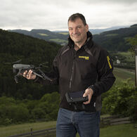 Thomas_Fleer Auf dem Bild sieht man einen Mann vor einer grünen Landschaft. In der Hand hält er eine Drohne. er trägt eine blaue Jean und eine schwarze Jacke, auf der ÖAMTC Fahrtechnik steht.