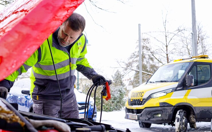 Ein ÖAMTC-Pannenhelfer überprüft im Winter den Motorraum eines Autos, während im Hintergrund ein ÖAMTC-Pannenfahrzeug am Straßenrand steht.