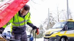 Ein ÖAMTC-Pannenhelfer überprüft im Winter den Motorraum eines Autos, während im Hintergrund ein ÖAMTC-Pannenfahrzeug am Straßenrand steht.