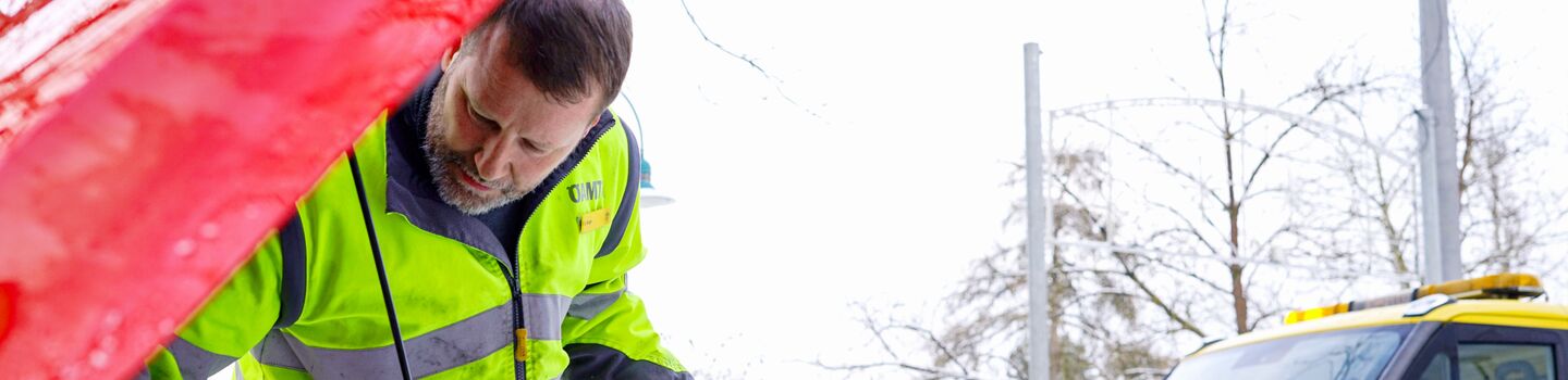 Ein ÖAMTC-Pannenhelfer überprüft im Winter den Motorraum eines Autos, während im Hintergrund ein ÖAMTC-Pannenfahrzeug am Straßenrand steht.
